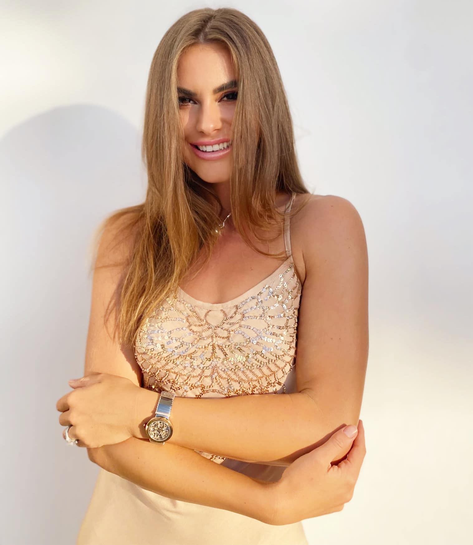 A woman with long brown hair smiles while wearing a beige dress adorned with sequins. She stands against a plain white background, arms crossed, and a silver watch on her left wrist—ready to help you find the perfect coach for women's coaching. coaching